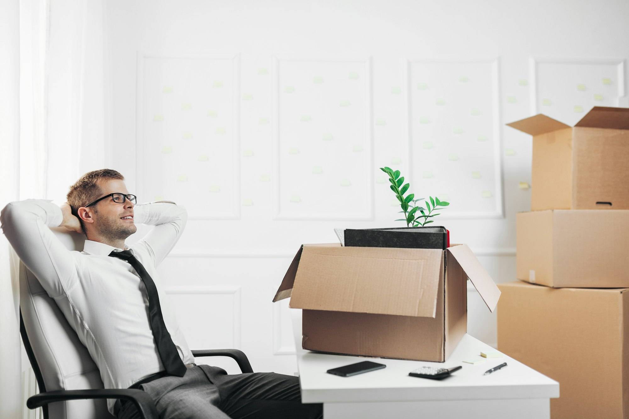 Homme d’affaires relaxé dans un bureau en déménagement, assis sur chaise, cartons empilés et plante sur le bureau. Démarches administratives déménagement bureau évoqué par le contexte.