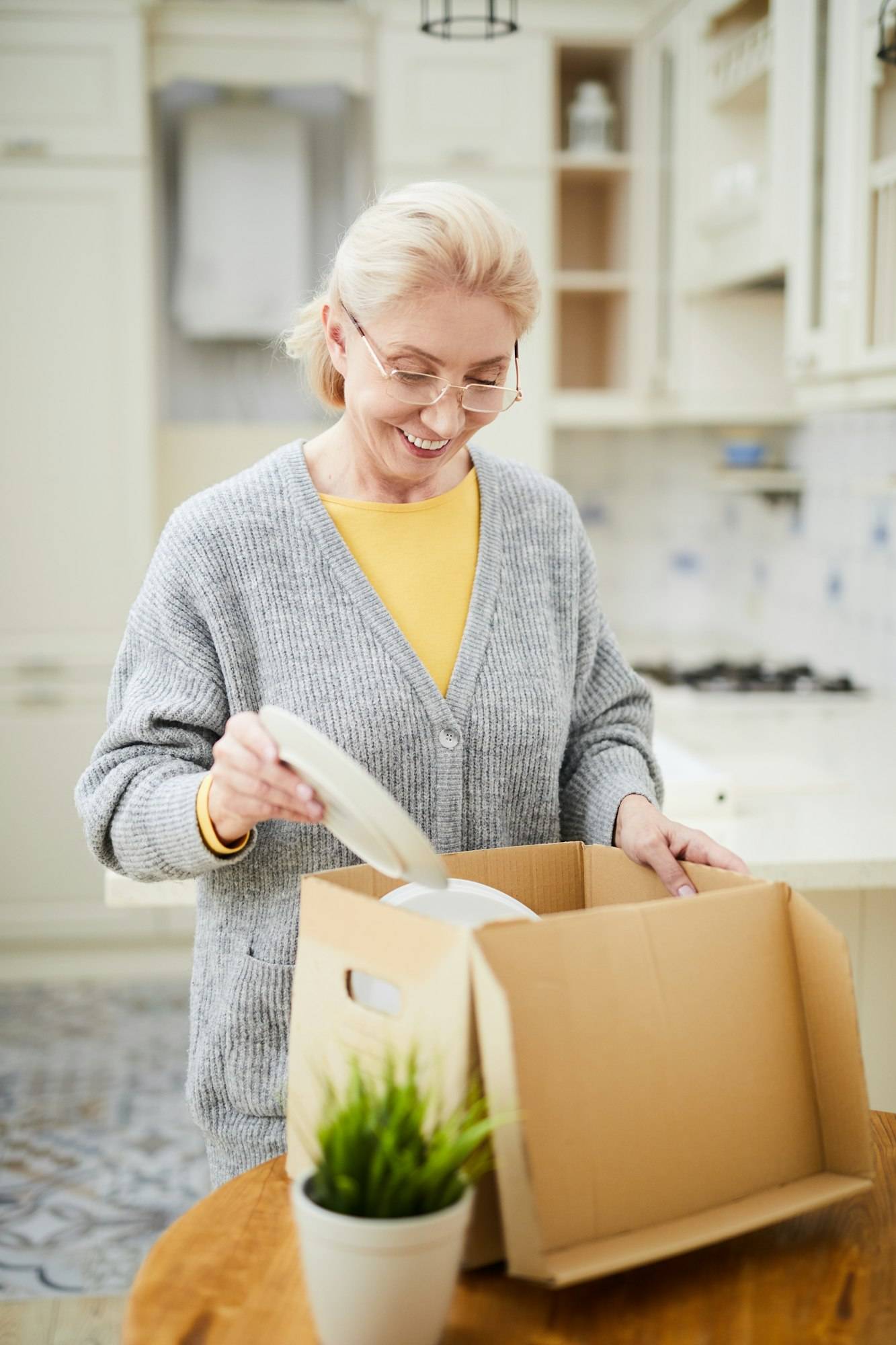 Personne âgée souriante qui emballe des objets dans une boîte en carton dans une cuisine, avec plante verte sur la table. Délai déménagement bureau professionnel intégré indirectement.