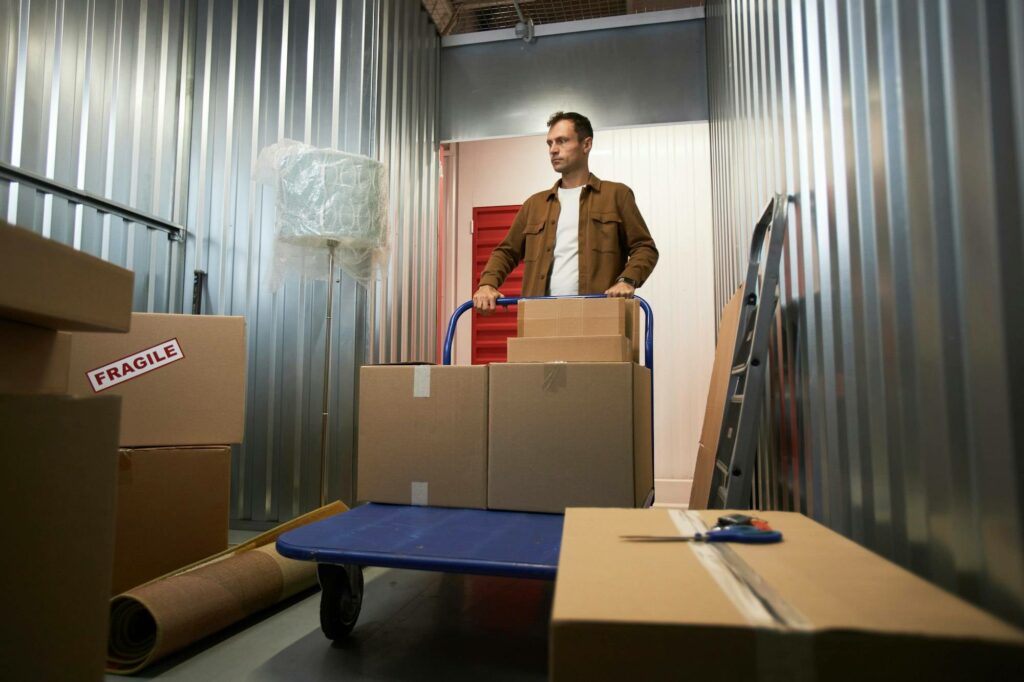 Homme déplaçant une valise de cartons sur un chariot dans un espace de rangement, entouré de boîtes et d’un étiquetage fragile, prêt pour un tri rapide. 14 choses à jeter en un jour pour être mieux.