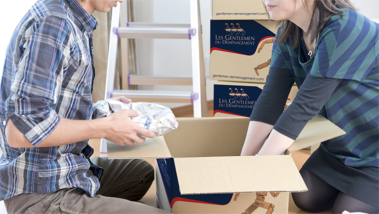 Deux personnes déballent des cartons dans une pièce remplie d’emballages et de boîtes.