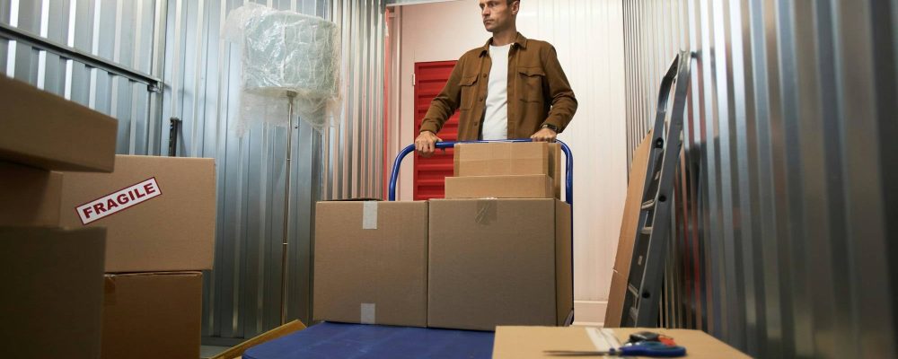 Homme déplaçant une valise de cartons sur un chariot dans un espace de rangement, entouré de boîtes et d’un étiquetage fragile, prêt pour un tri rapide. 14 choses à jeter en un jour pour être mieux.
